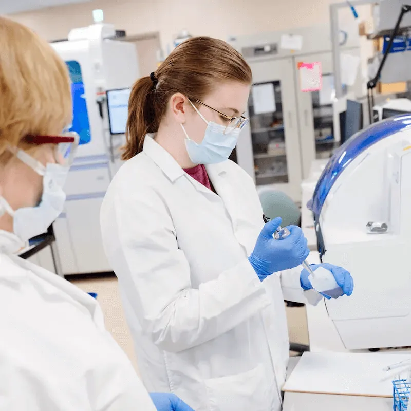 Two lab workers in masks and gloves preparing samples in a clinical laboratory.
