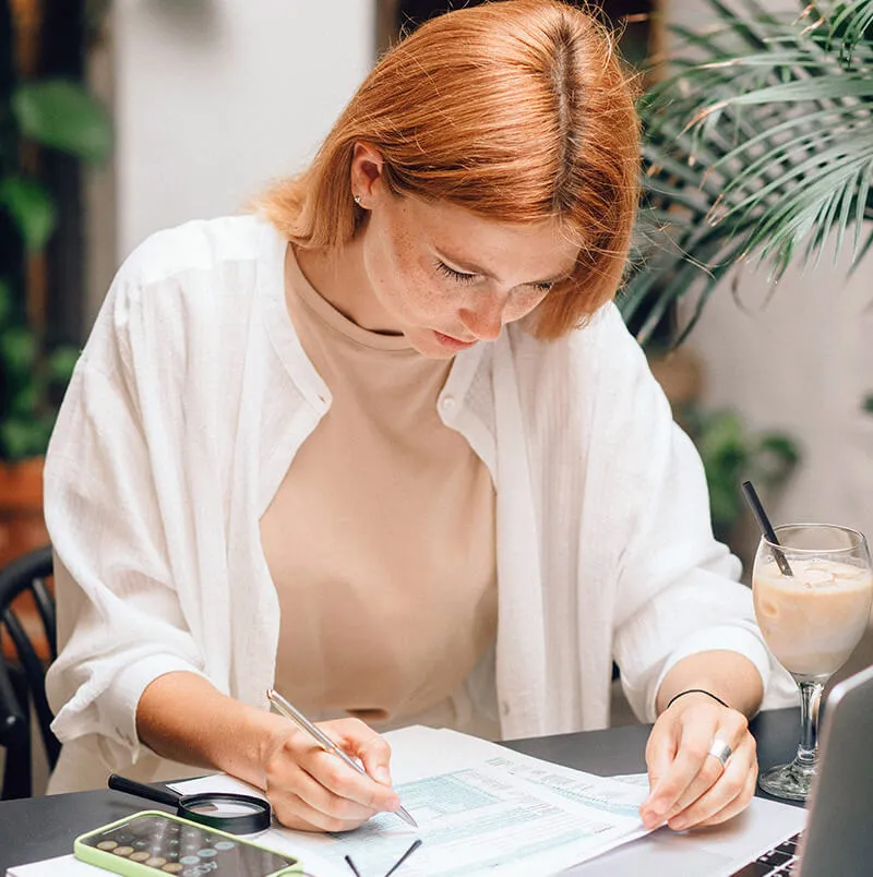 Person writing on a document at a desk with a laptop and drink.