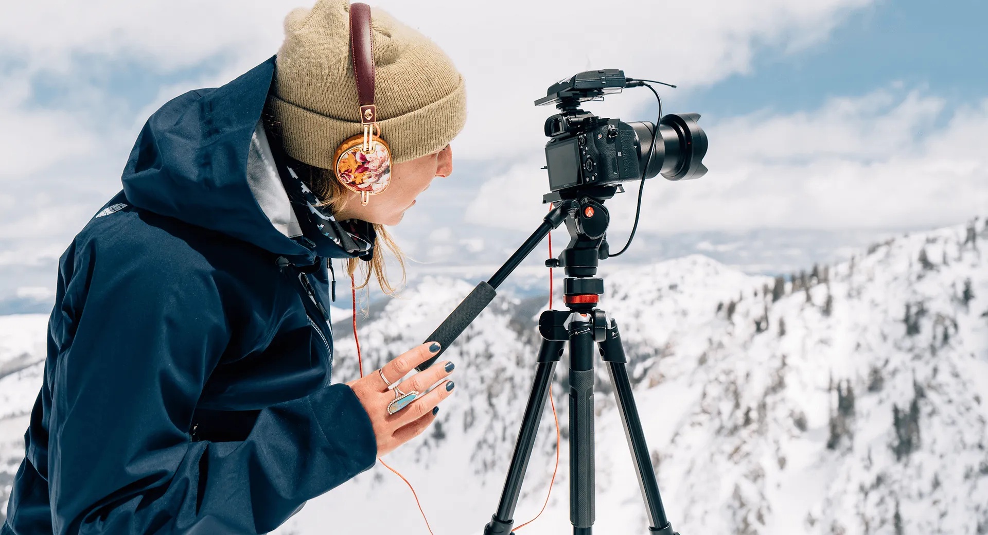Person in winter gear adjusting a camera on a tripod in a snowy mountain landscape.