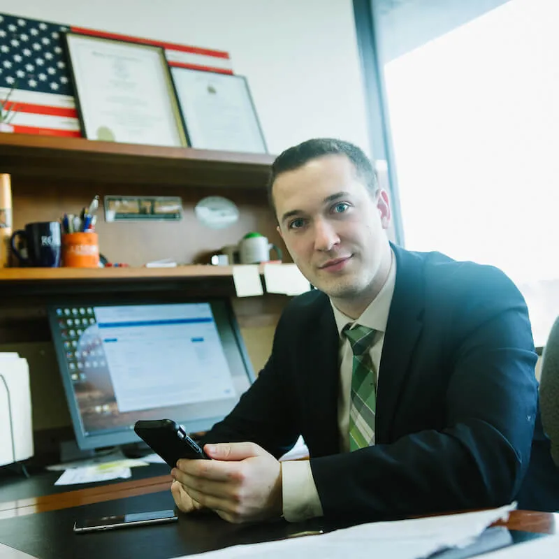 A person in a suit sits at an office desk, holding a phone, with a computer monitor and bookshelves in the background.