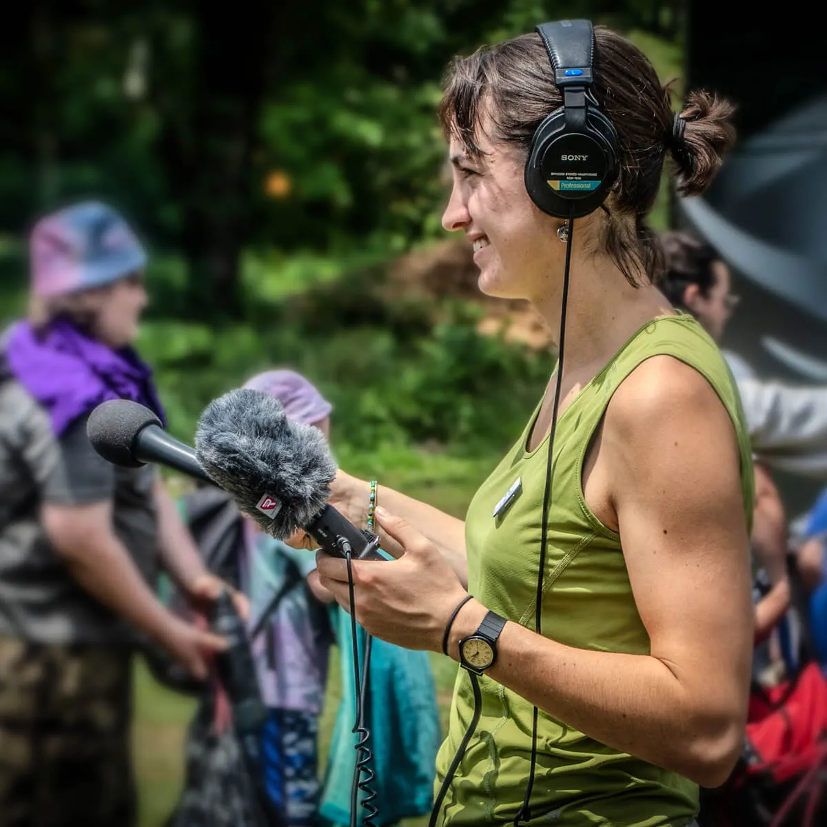 A person outdoors wearing headphones and holding a microphone with a windscreen while recording audio.