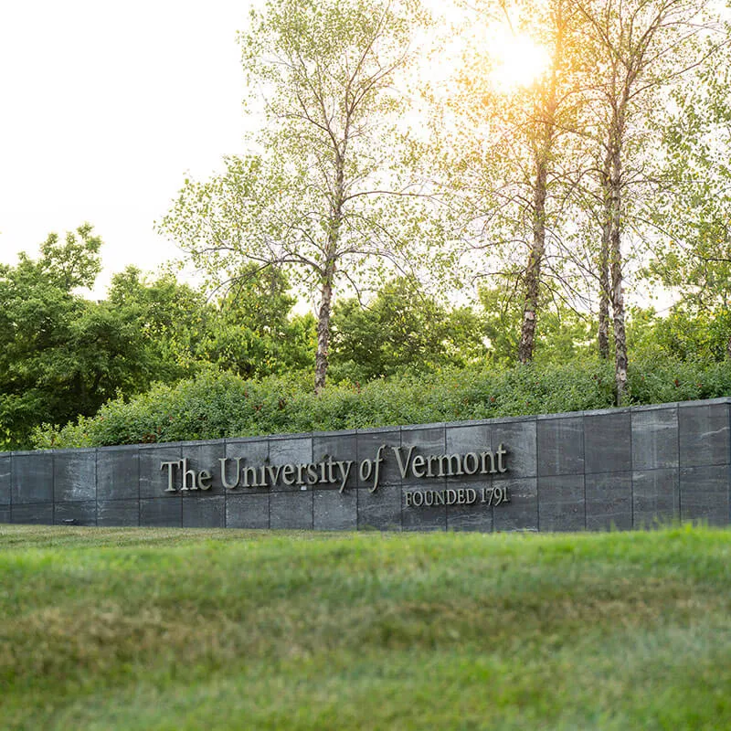 Stone wall sign reads The University of Vermont with sun light peaking through trees in background.