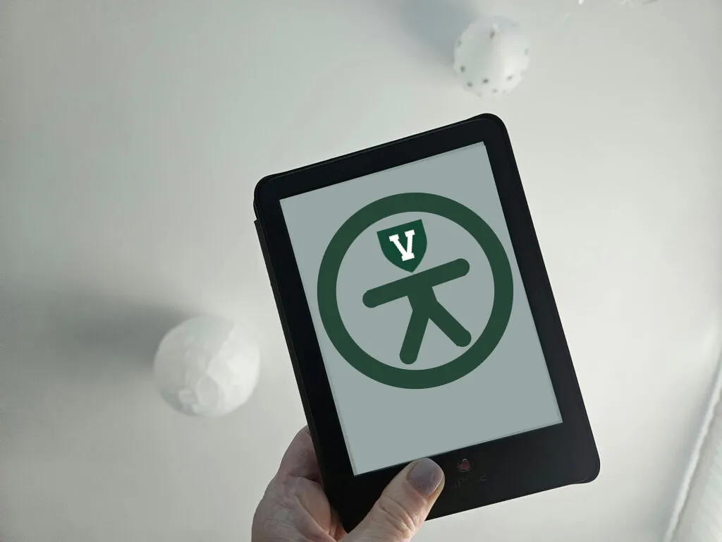 Hand holding a Kindle-style tablet. On screen is the icon for accessibility with a UVM V for a head