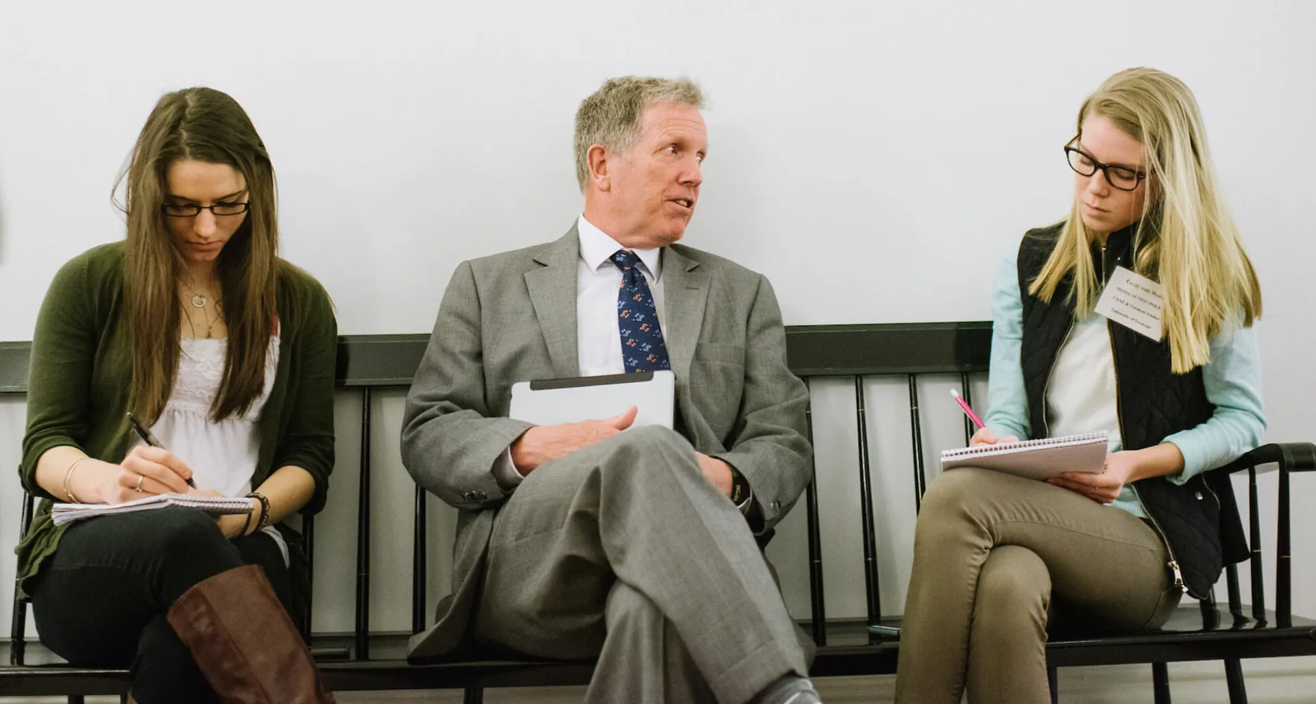 A person in a suit talks with two individuals seated beside them on a bench