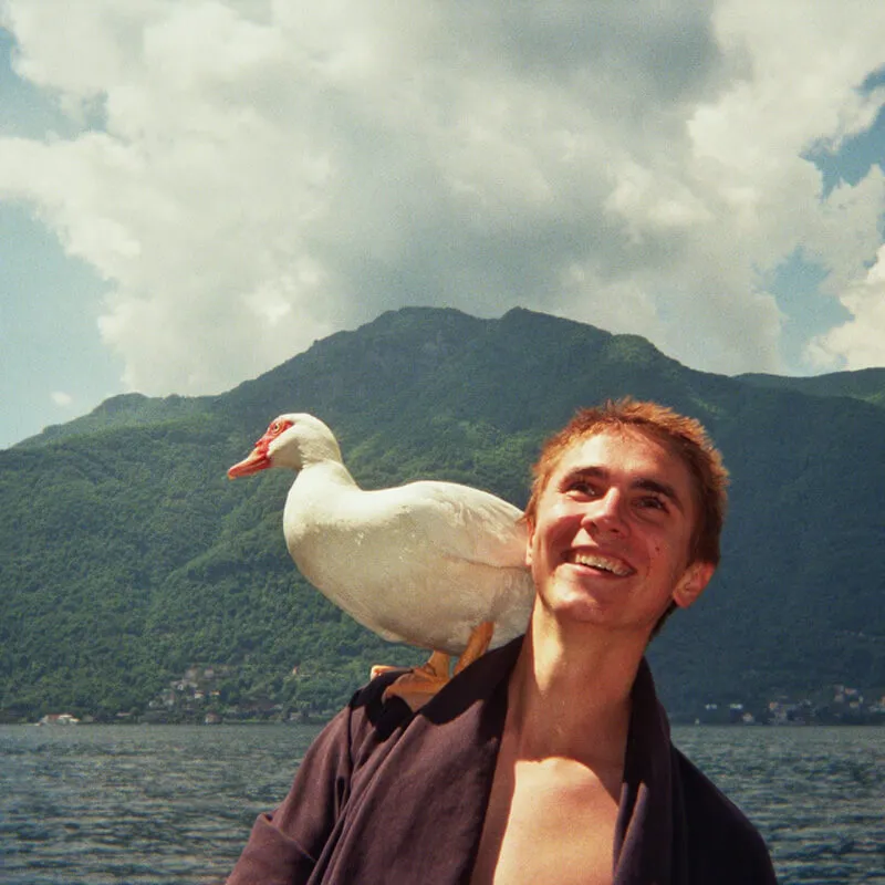 tudent smiling with a white duck perched on their shoulder beside a lake and green mountains.