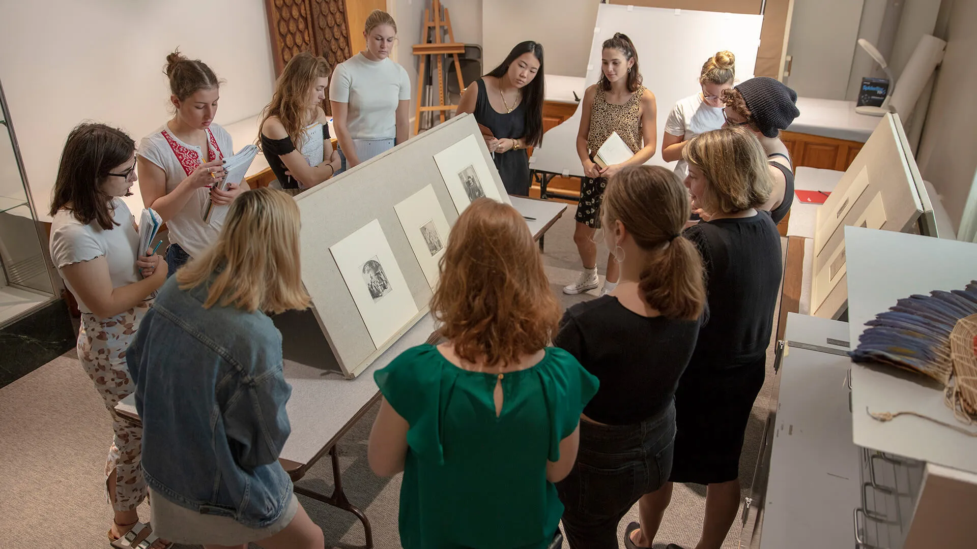 A group gathers around displayed artwork in a studio, looking closely and discussing the pieces.