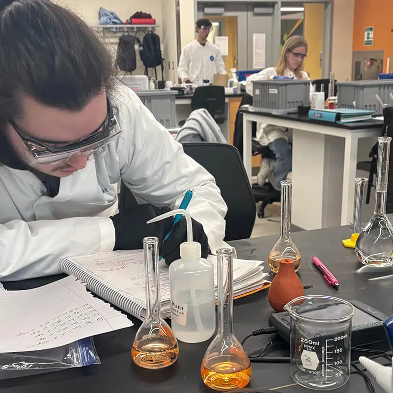 Student in a chemistry lab records notes while working with glassware at a lab bench.