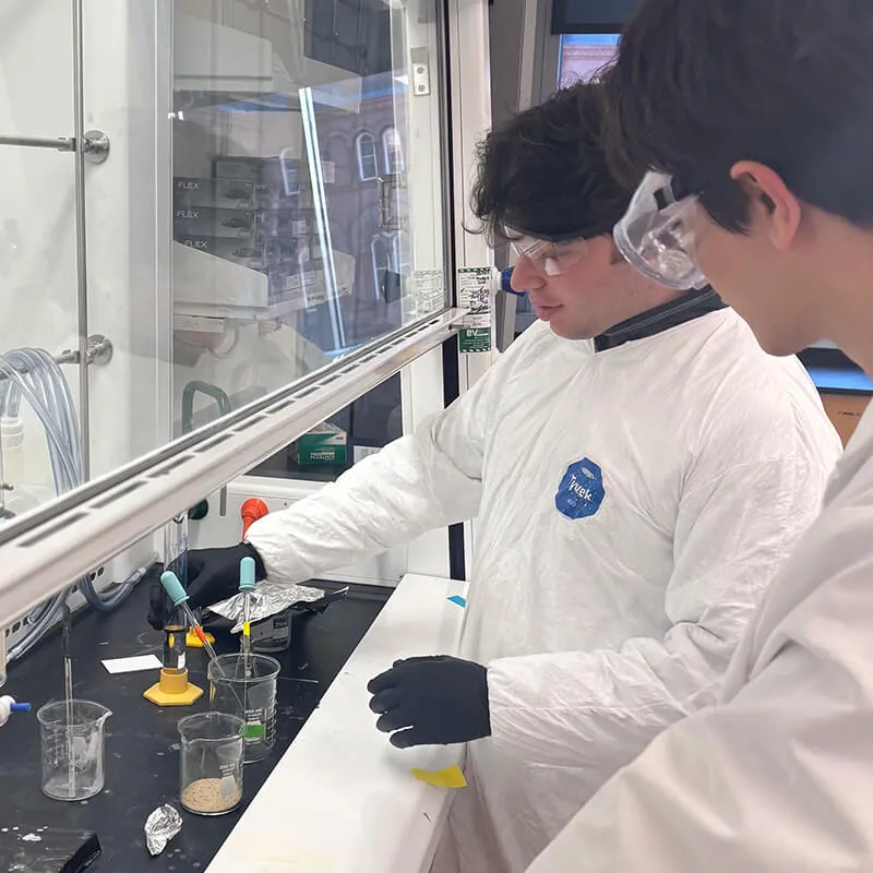 Two students wearing lab coats and safety goggles conduct an experiment together at a chemistry lab bench.