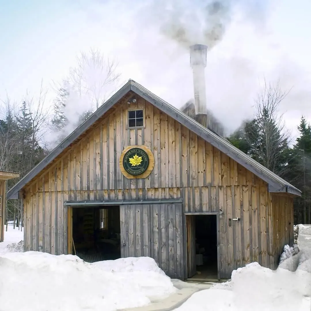 a large wooden sugarhouse in snow with a steaming chimney pipe