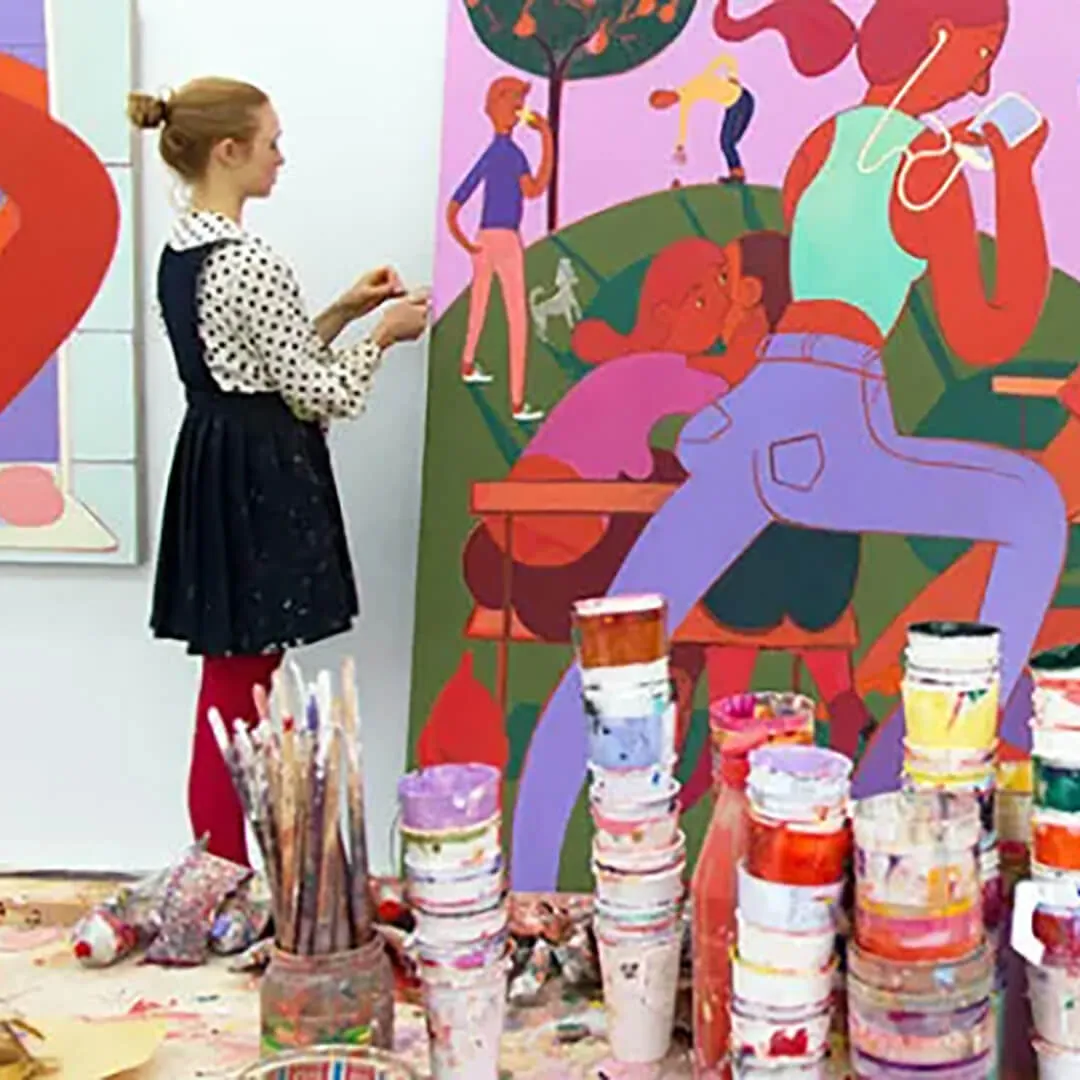 A person works in an art studio beside a large, colorful painting, with paint containers and brushes in the foreground.