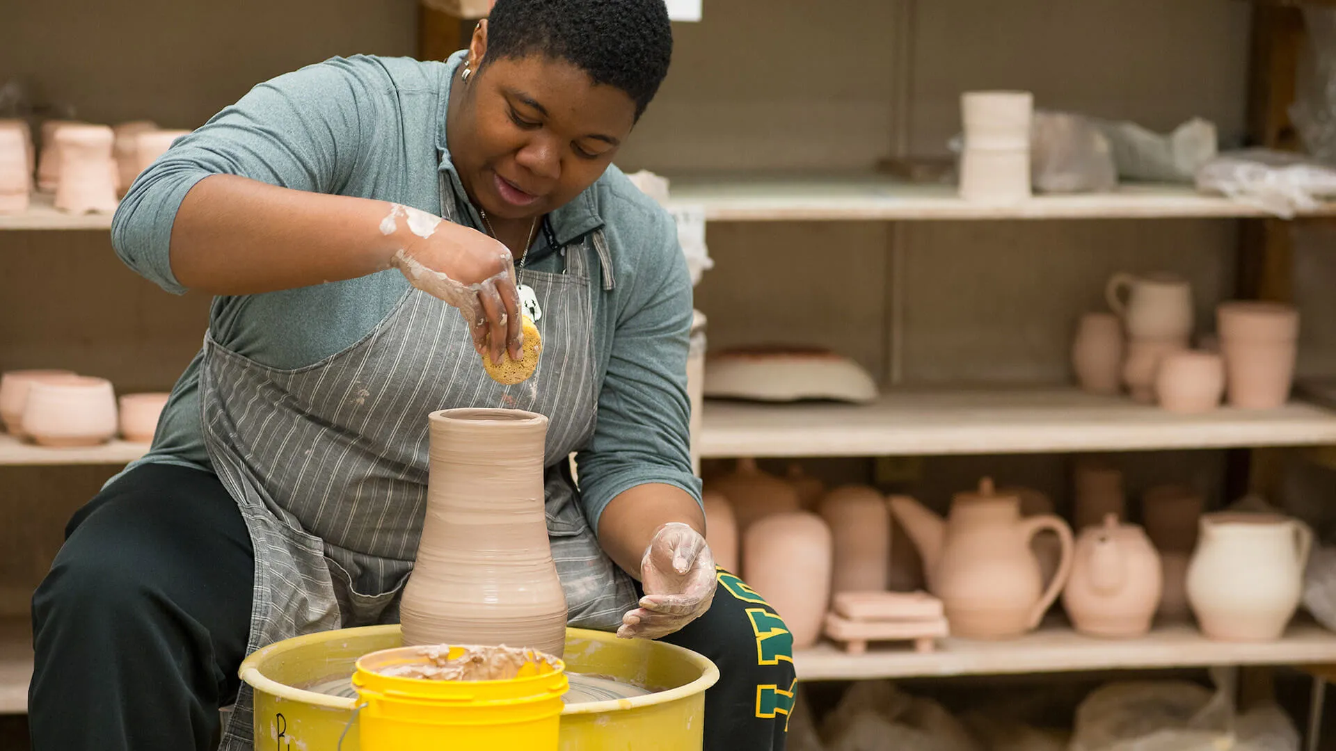 Person shapes a clay vessel on a pottery wheel in a studio.