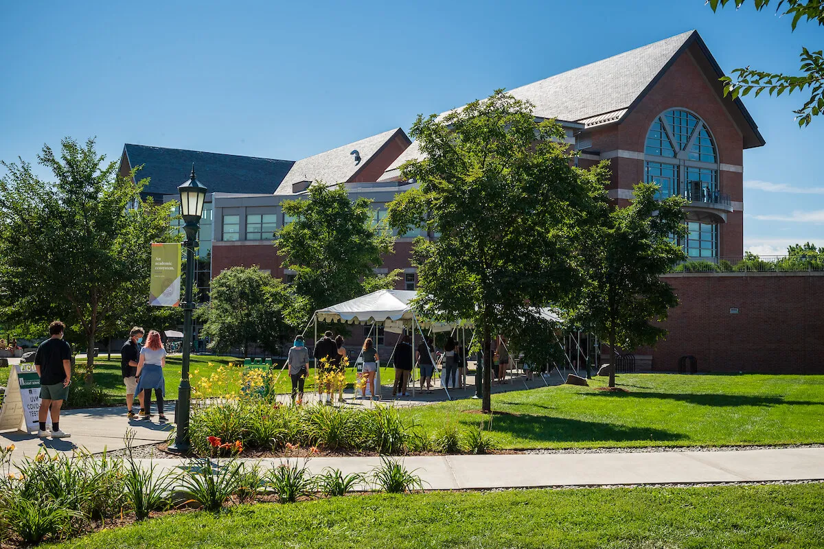 “Students walk across a sunny college campus with green lawns and trees, passing a white event tent in front of a large brick academic building.