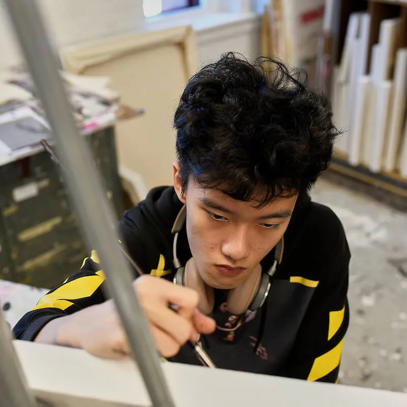 Person concentrates while painting or drawing at a worktable in a studio.