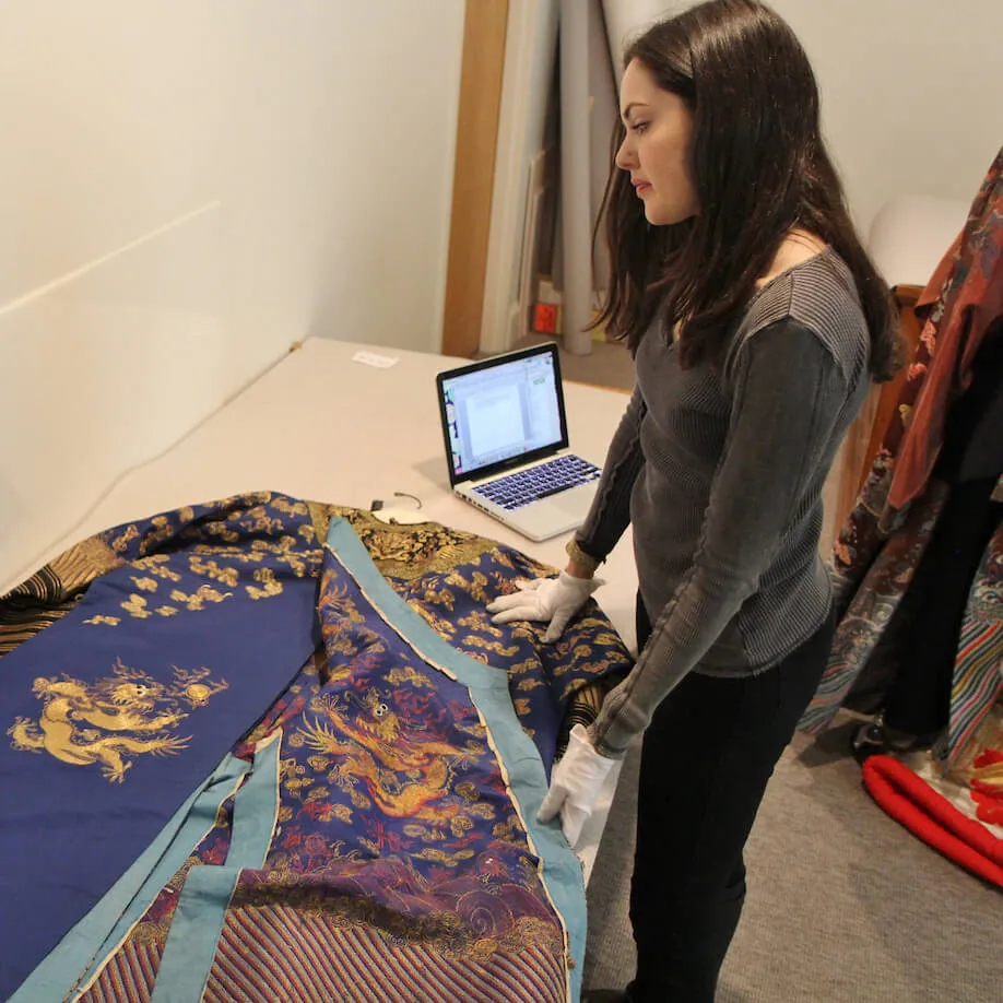 Person wearing gloves examines an ornate embroidered garment laid out on a table beside a laptop.