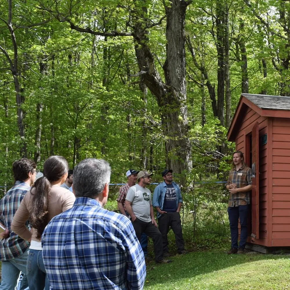 a man speaking to a tour group outside a red outbuilding in summertime