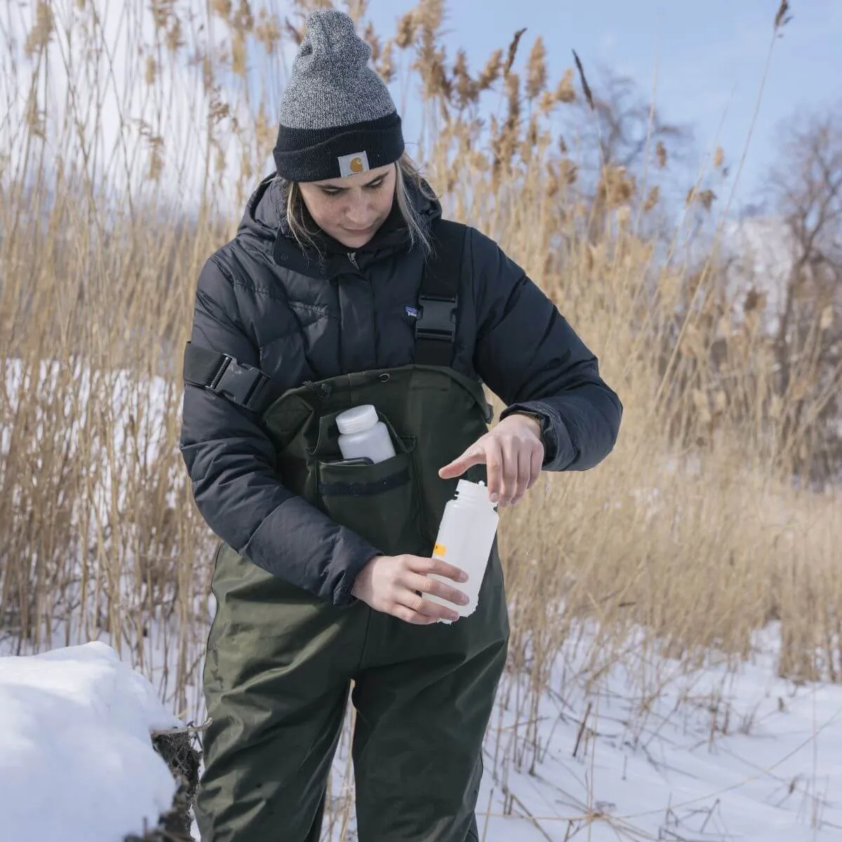 A student collecting samples outside on a snowy day, they are wearing warm-weather gear and screwing the lid on a bottle