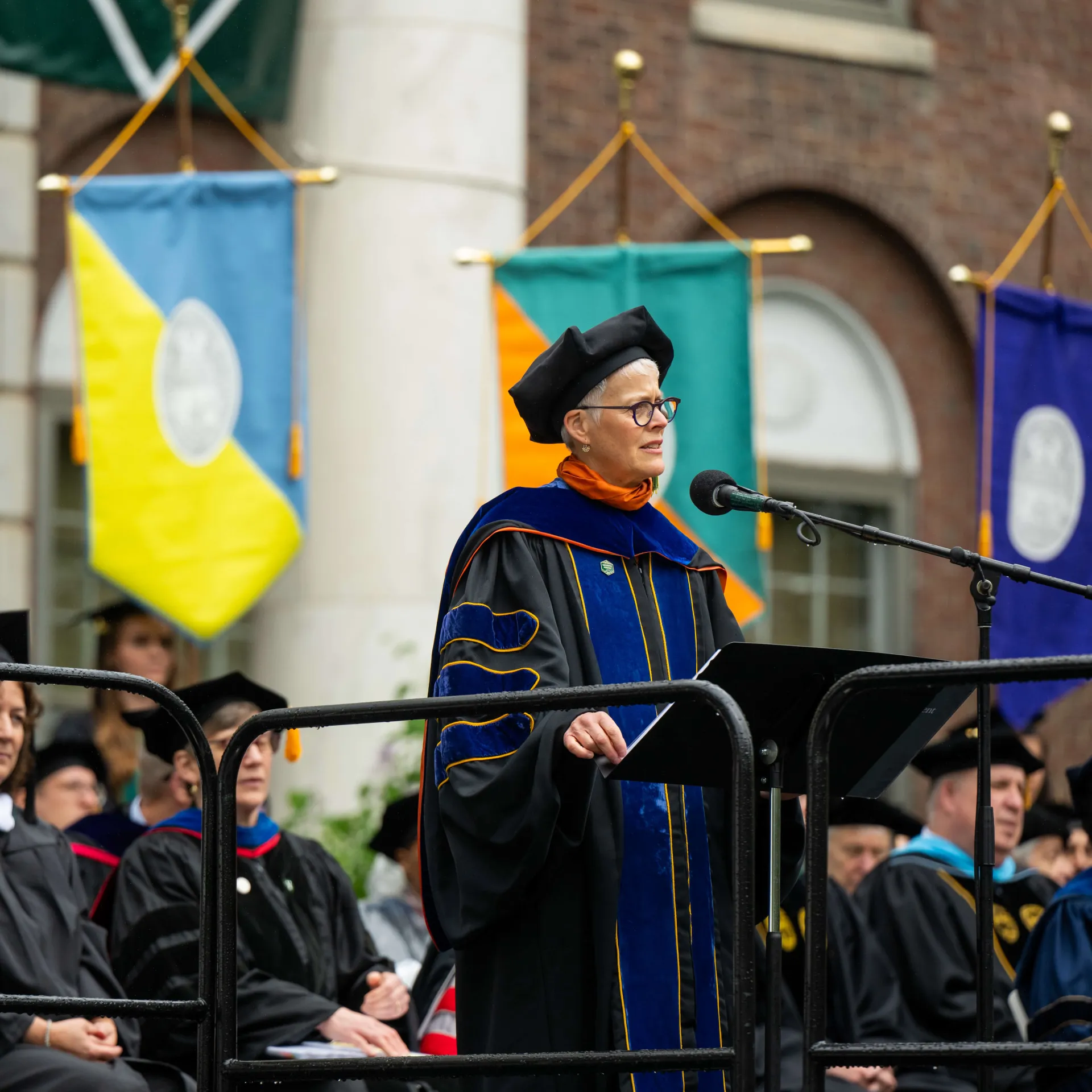 A woman with light skin and short grey hair, wearing glasses and academic regalia, stands at a microphone on stage during commencement ceremony