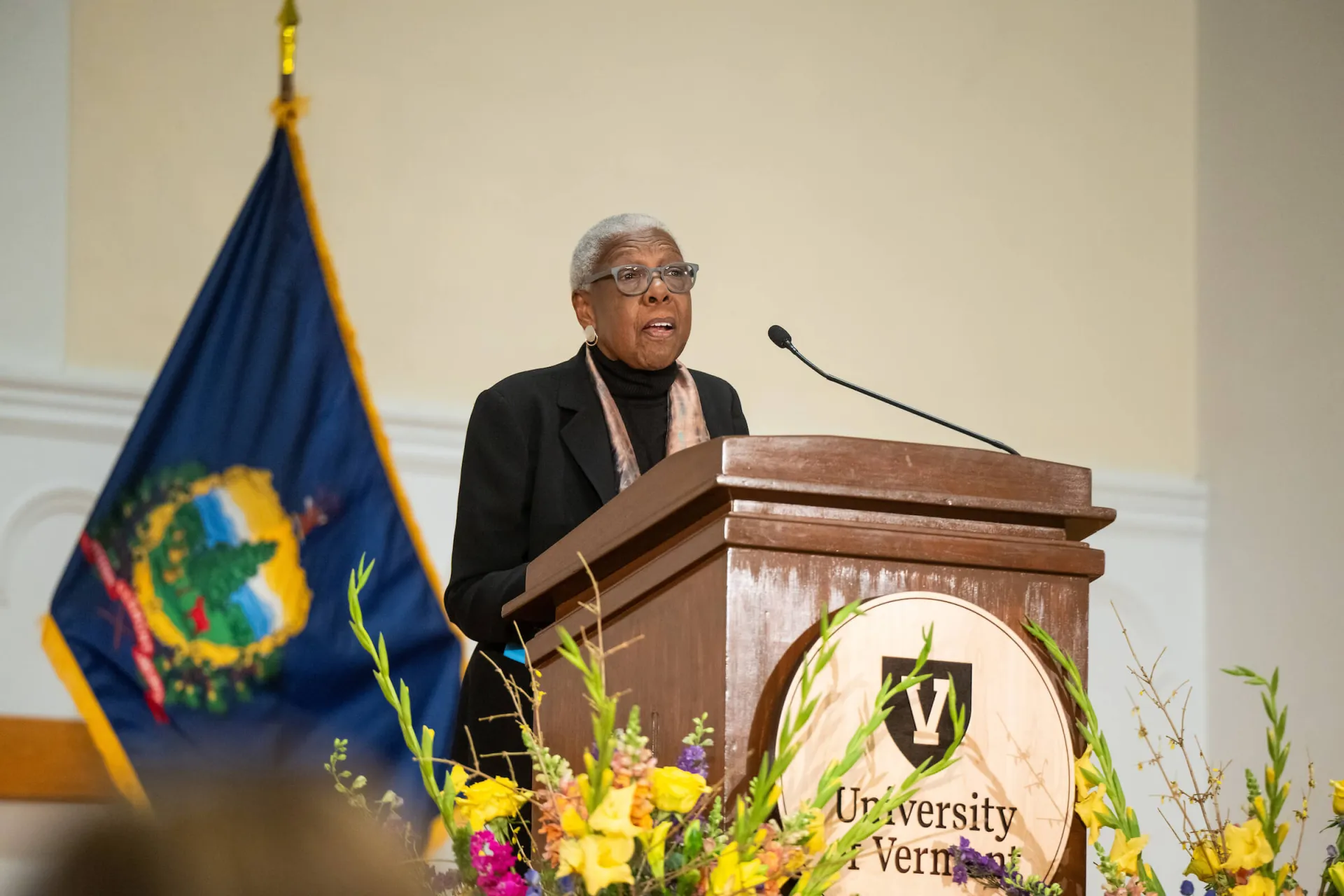 2026 MLK Keynote Speaker Rosalyn Pelles speaking behind a UVM Podium with the Vermont State Flag behind her. 