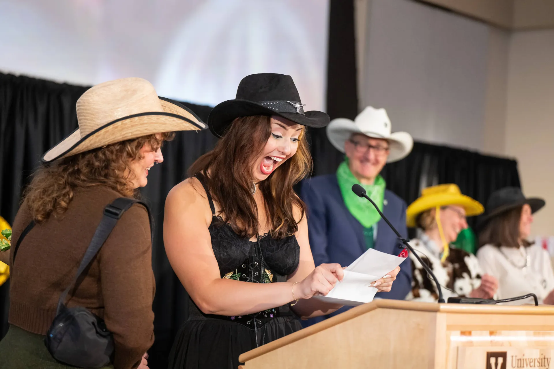 People in Western-style hats stand at a podium; one person at the front looks at a paper while others stand behind onstage.