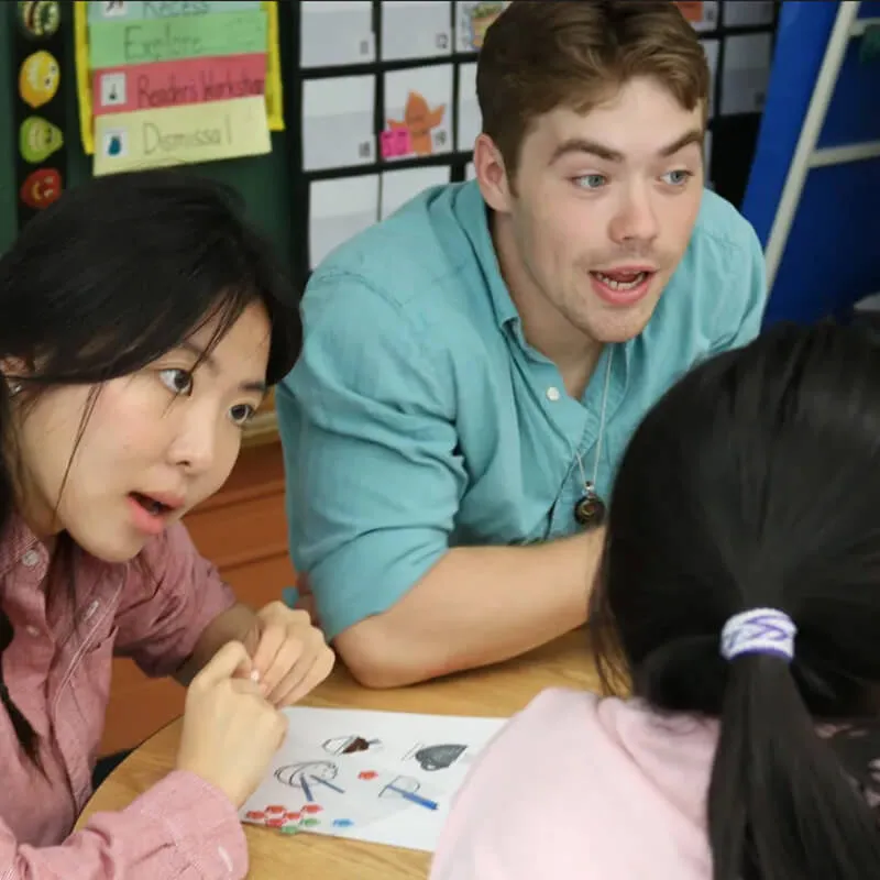 Two adults helping a child practice Mandarin at a classroom table.