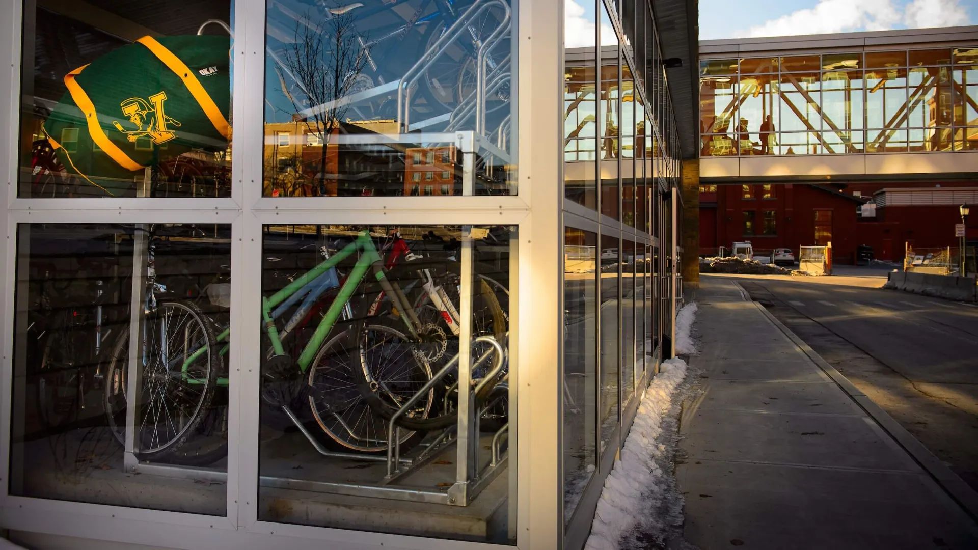 inside a building with glass walls, bicycles are stored. there is a small amount of snow on the sidewalk outside.
