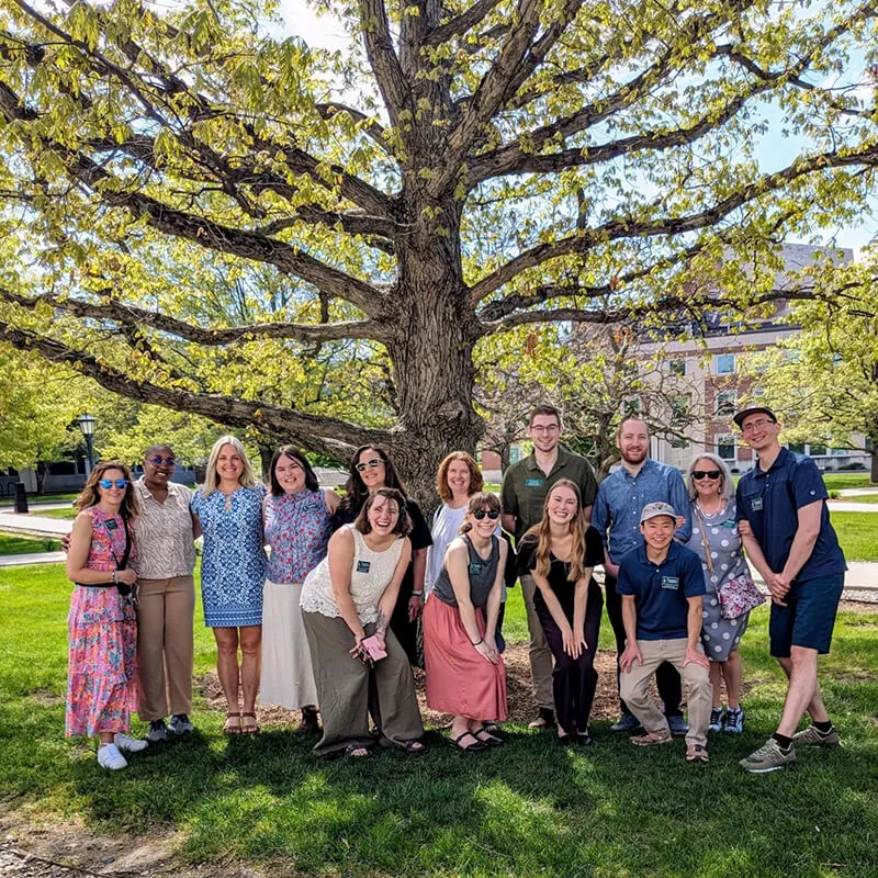 Group of people posing under a large tree on a sunny campus lawn.