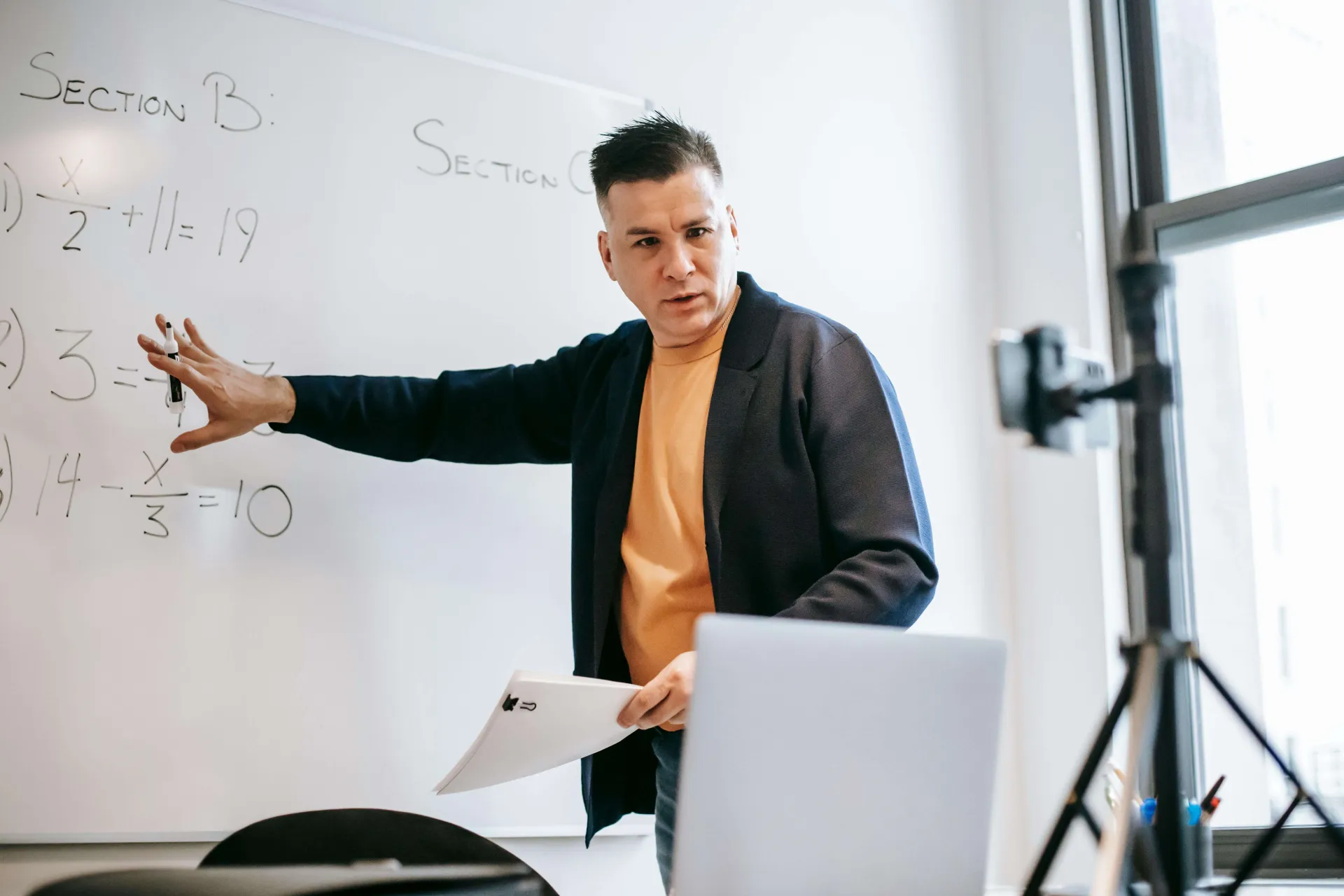 A Latinx professor gestures to an equation on a whiteboard while looking into a camera