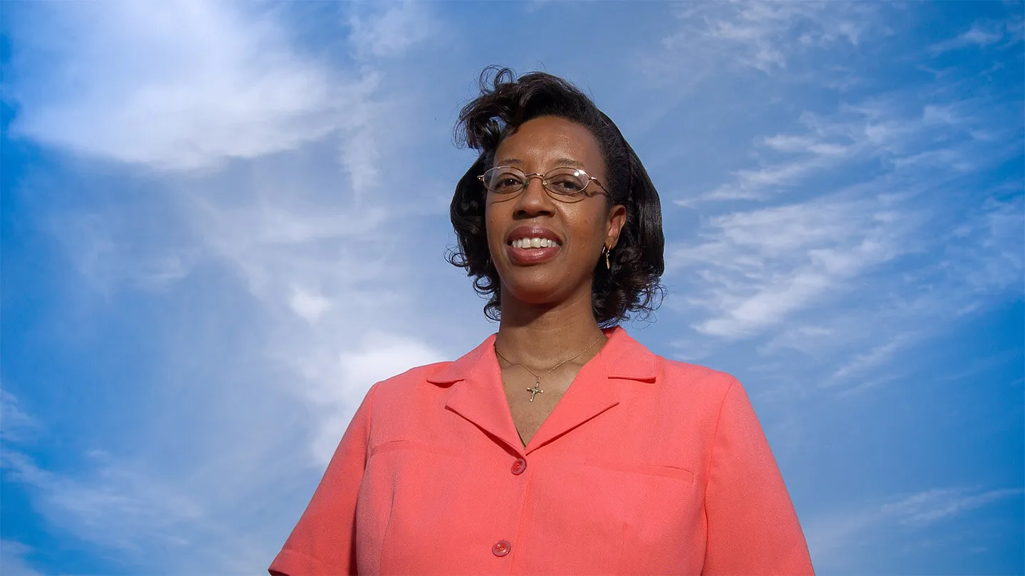 woman in pink shirt and glasses smiling with sky and clouds in background