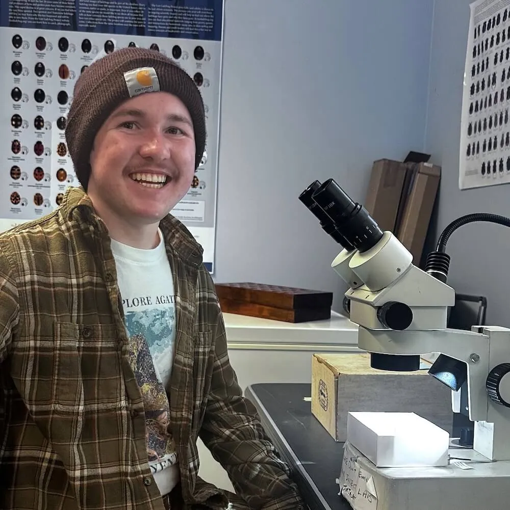 A young man in a plaid shirt sits at a table with a microscope, smiling, with beetle posters in the background