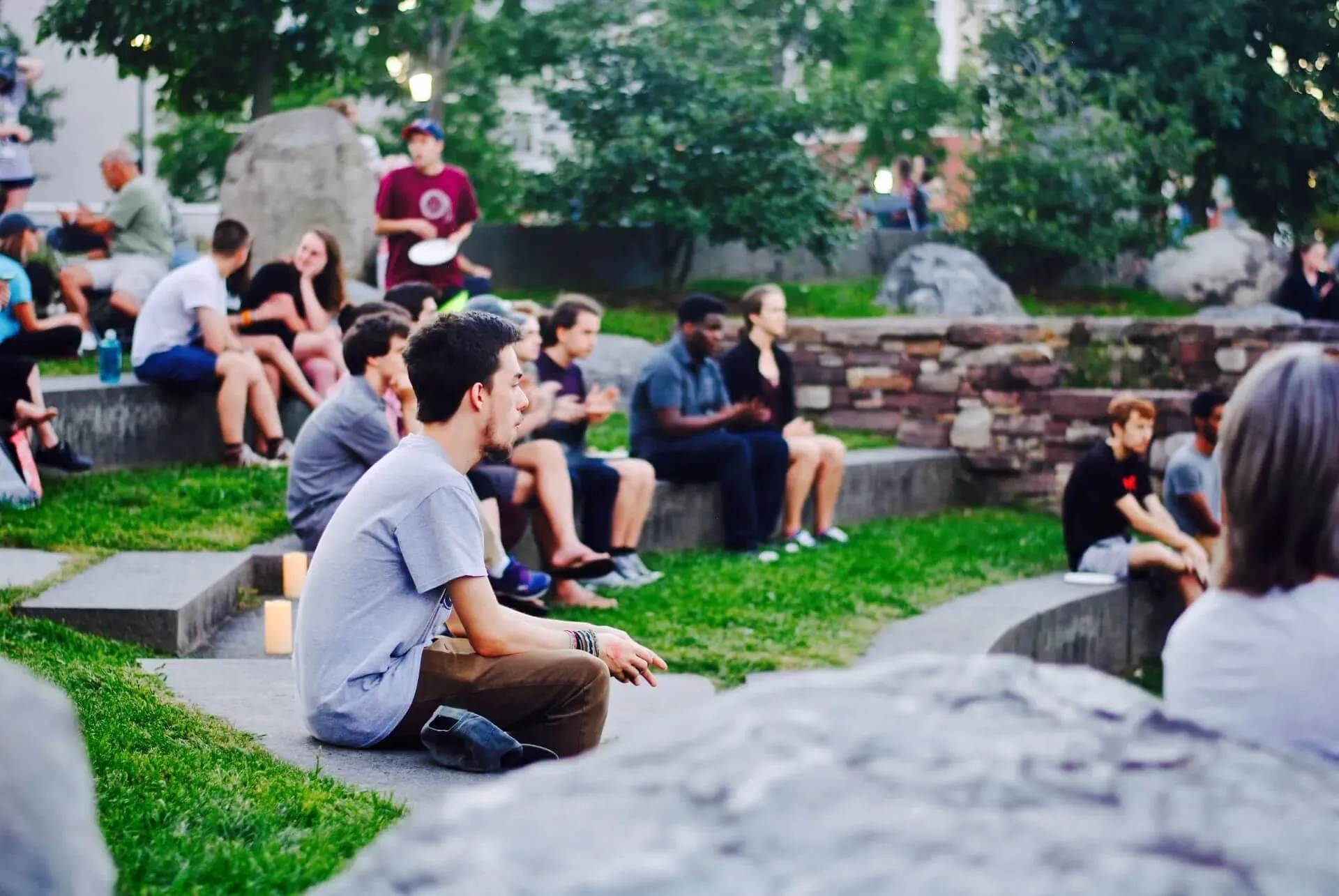 A group of students seated on the ground. They are setting on man-made stone ledges that are build like curved steps with a large patch of grass between each ledge where students are resting their legs. There is a student centered in the middle of the image.  