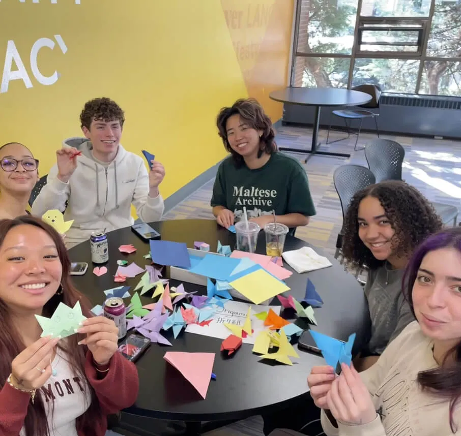 Six young adults smiling around a table, folding colorful origami with paper and drinks in a bright room.