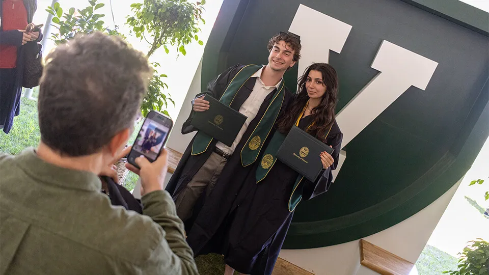 Graduates get their photo taken in front of the UVM shield at the Grossman School of Business commmencement