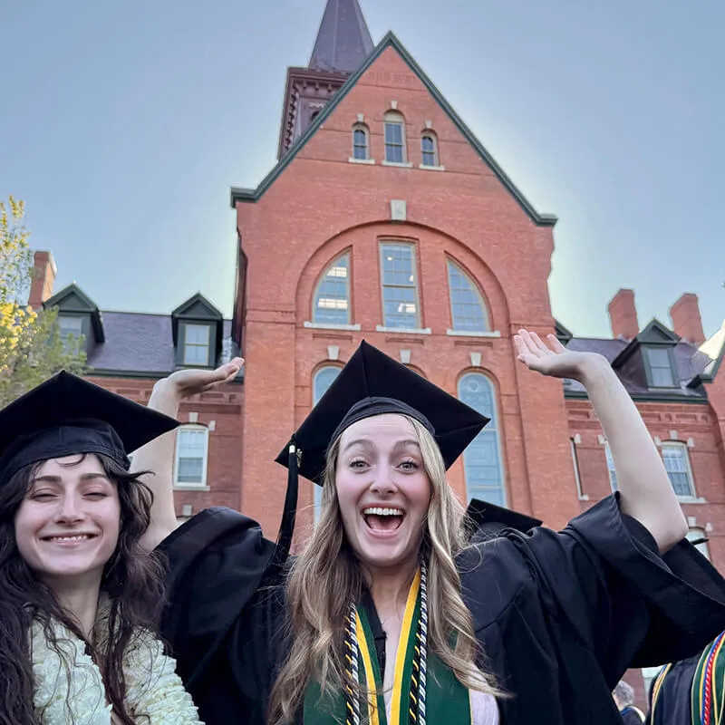 Two female students in black academic regalis (cap and gown) stand in front of red brick building (Old Mill). Both graduates are smiling. Student in the center is raising their arms in celebration. 