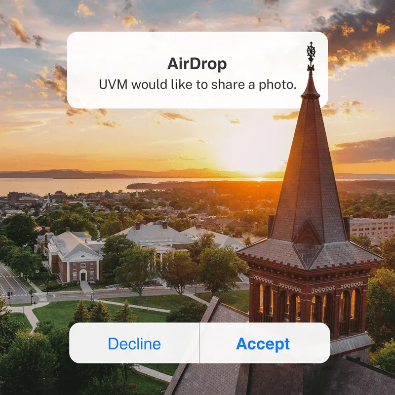 Sunset overlooking UVM's campus, Burlington, Lake Champlain and Adirondack Mountains. UVM's Old Mill tower overlays a message that reads, "Air Drop, UVM would liks eot share a photo. Decline or Accept." Accept is bolded. 