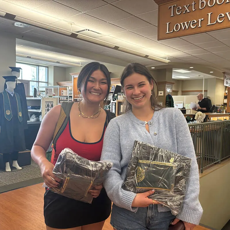 Two female students stand in the bookstore smiling holding their graduation cap, gown and stole.