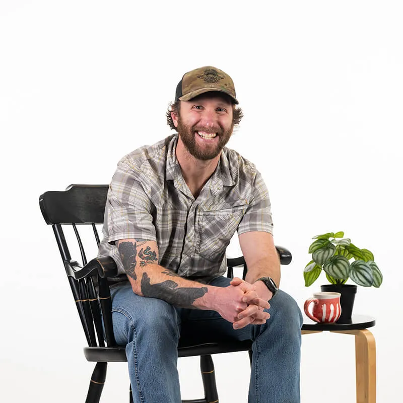 Male student wearing baseball cap, grey flannel shirt and blue jeans smiles while sitting in a chair leaning forward on knees. Backgroung is white. A small table with a green plant a coffee mug are next to the chair. 