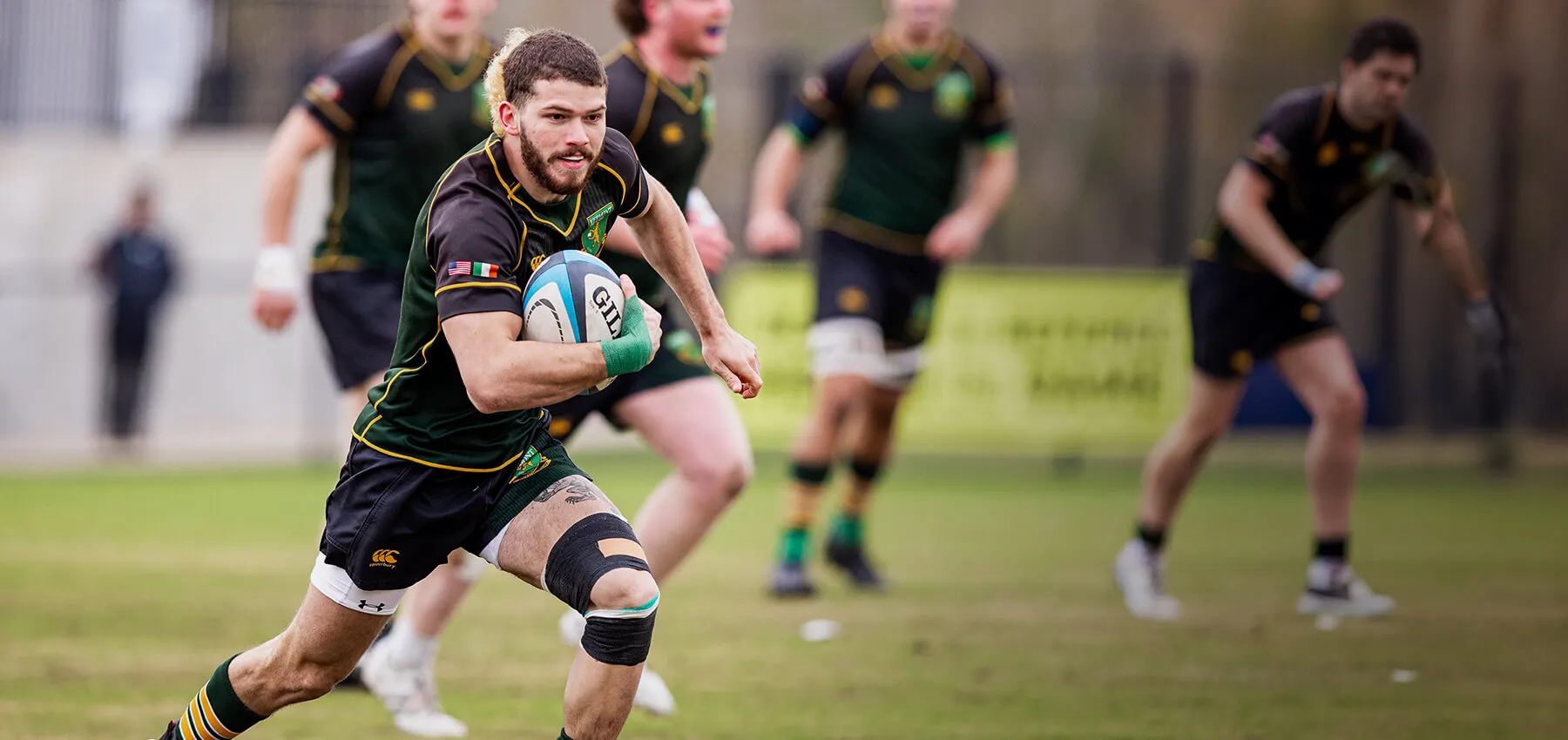 a mens rugby player holds a ball while sprinting across a field