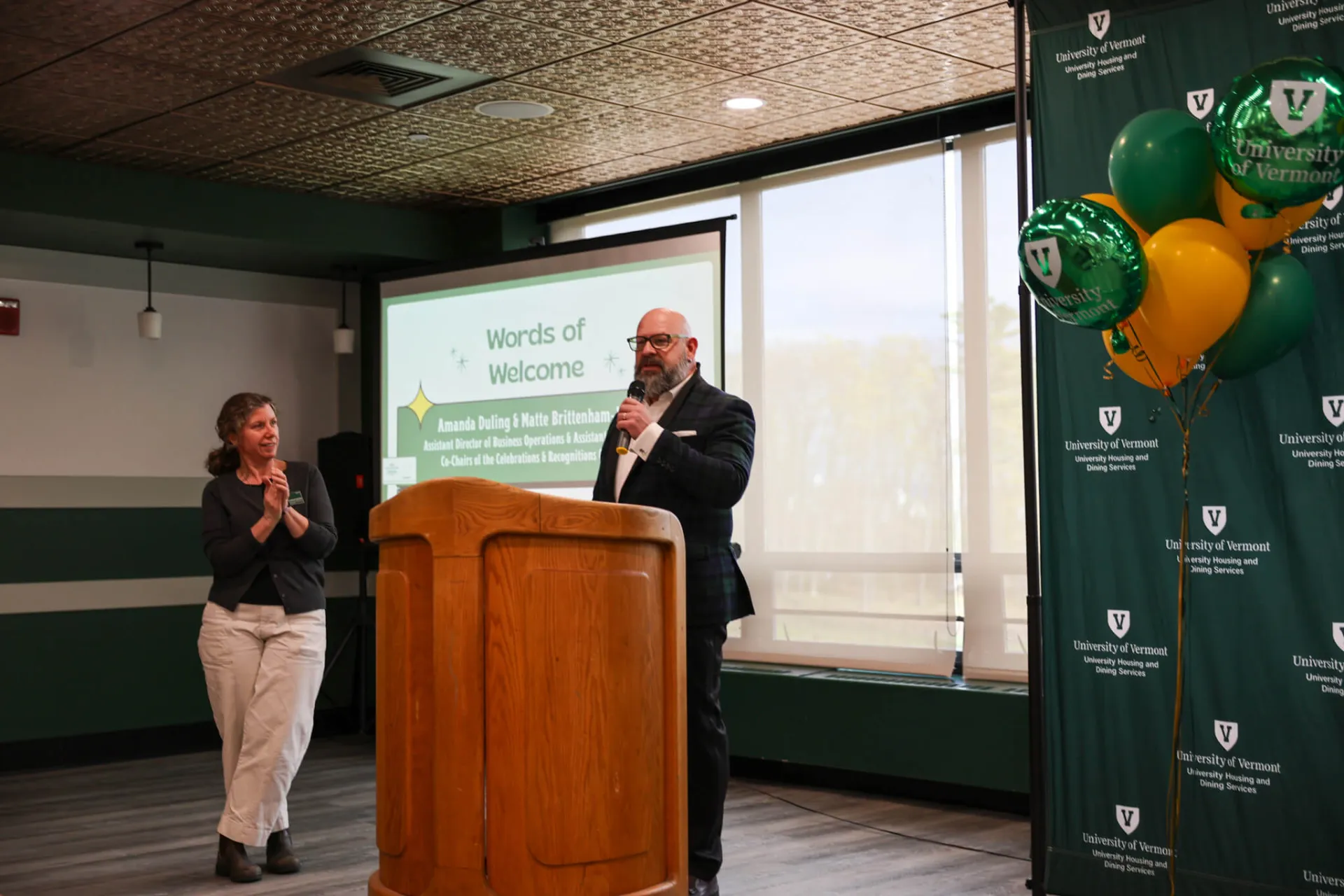 Two figures stand near a podium, one holds a microphone and is speaking. There is an UVM Housing and Dining Backdrop