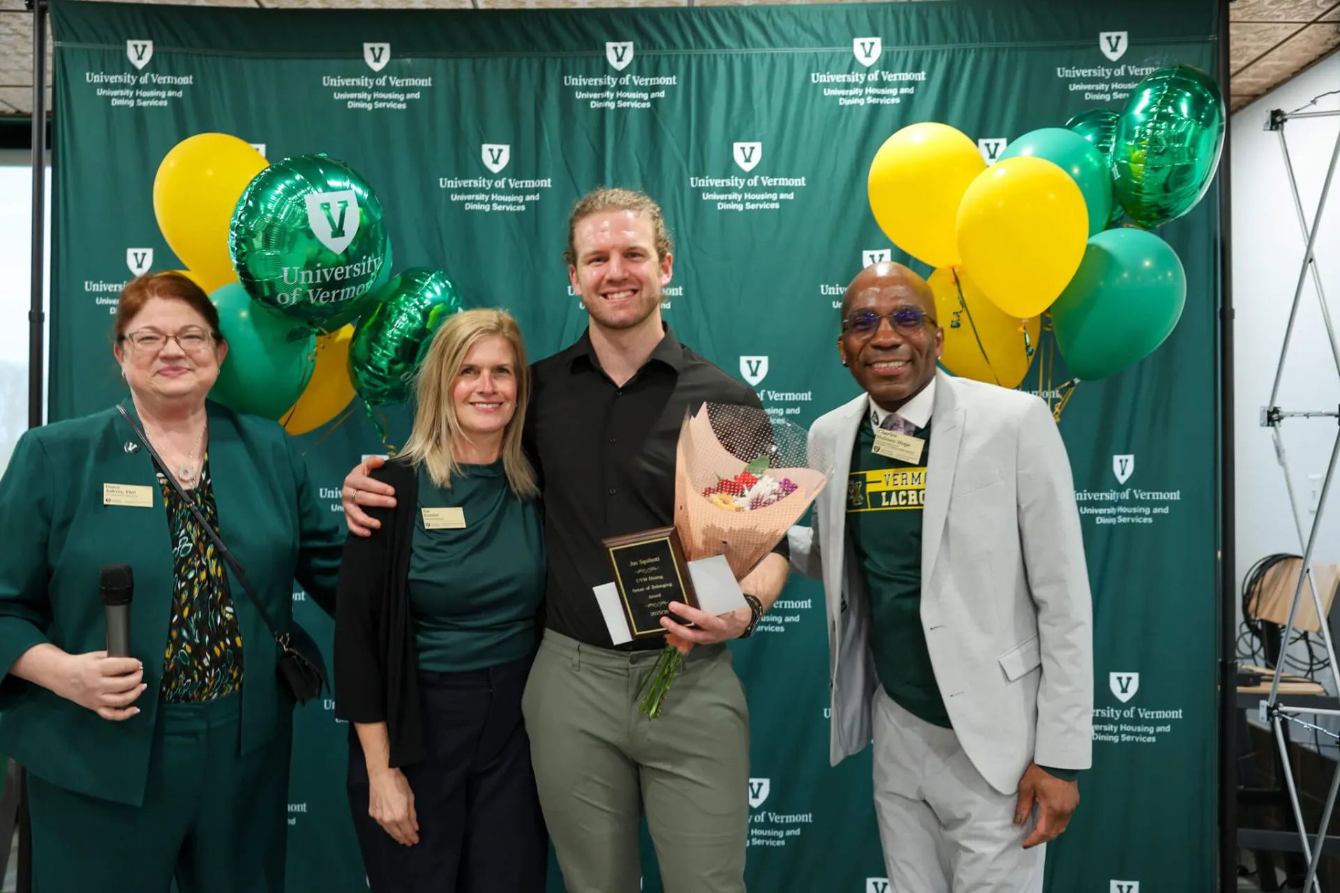 Four individuals stand in front of the Housing and Dining backdrop. One holds an award.