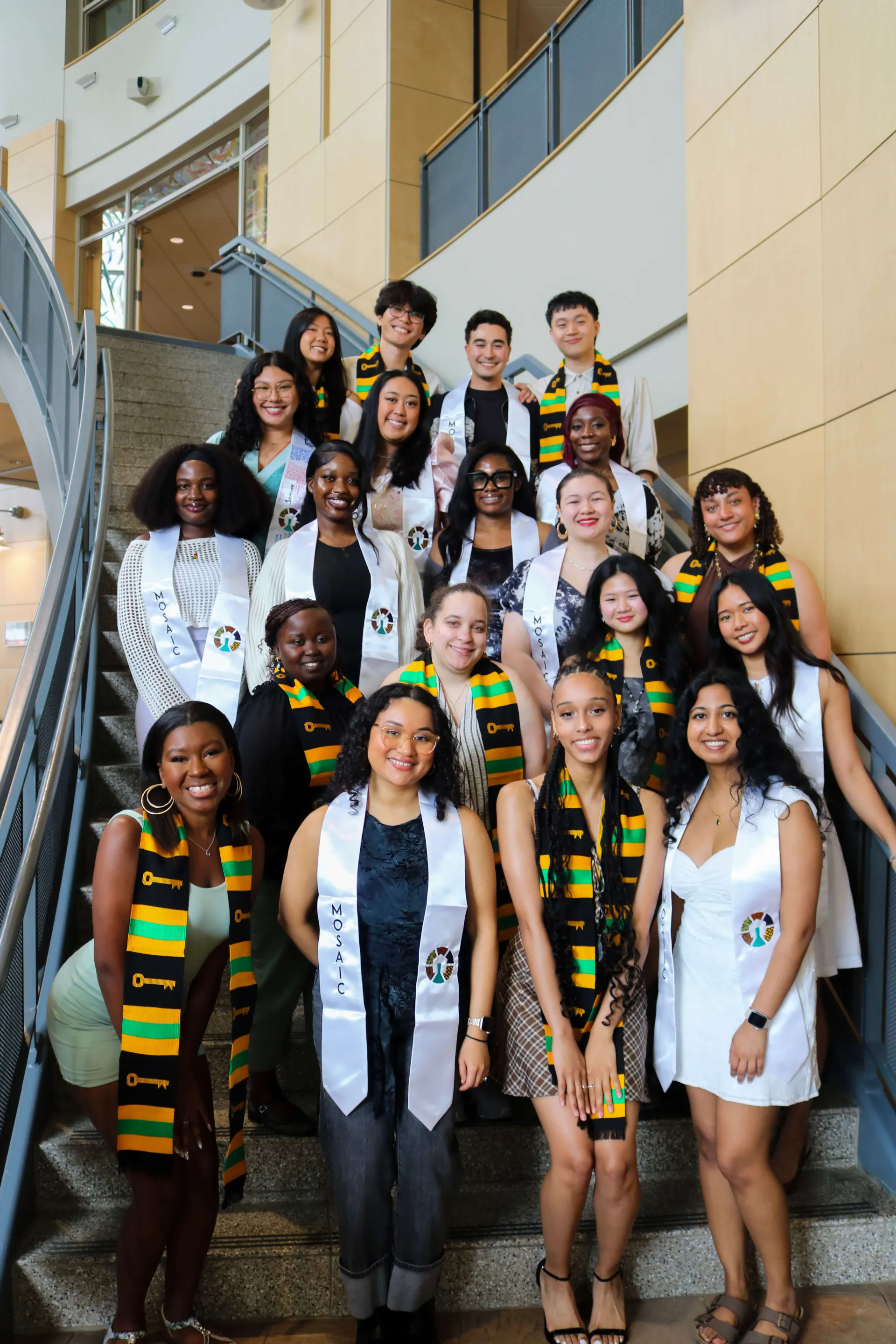 A group of graduating students from the Mosaic Intercultural Center stands together on the steps of the Davis Center, wearing Mosaic graduation stoles and smiling as they celebrate their achievement.