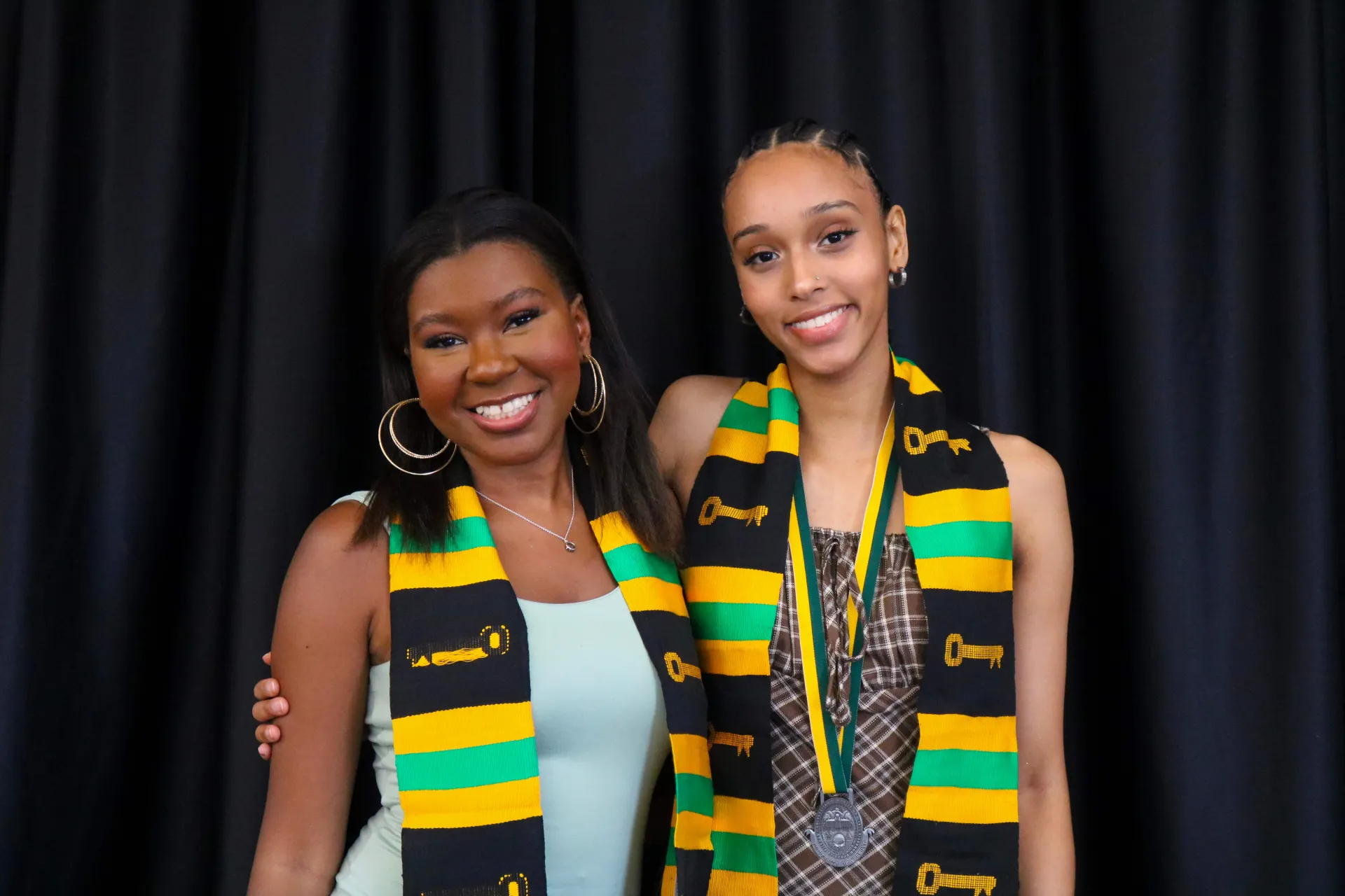 Two students smiling and posing against a black background and they are wearing Kente graduation stole 