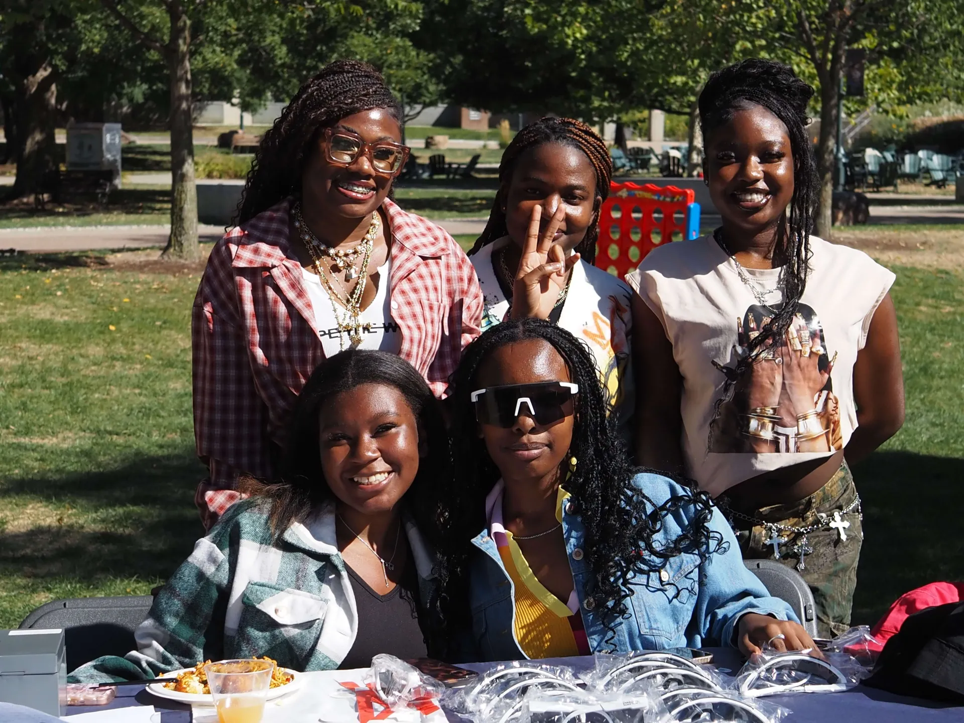 A group of students some are siting at a table with sunglasses and food and other are standing behind them and they are smiling 