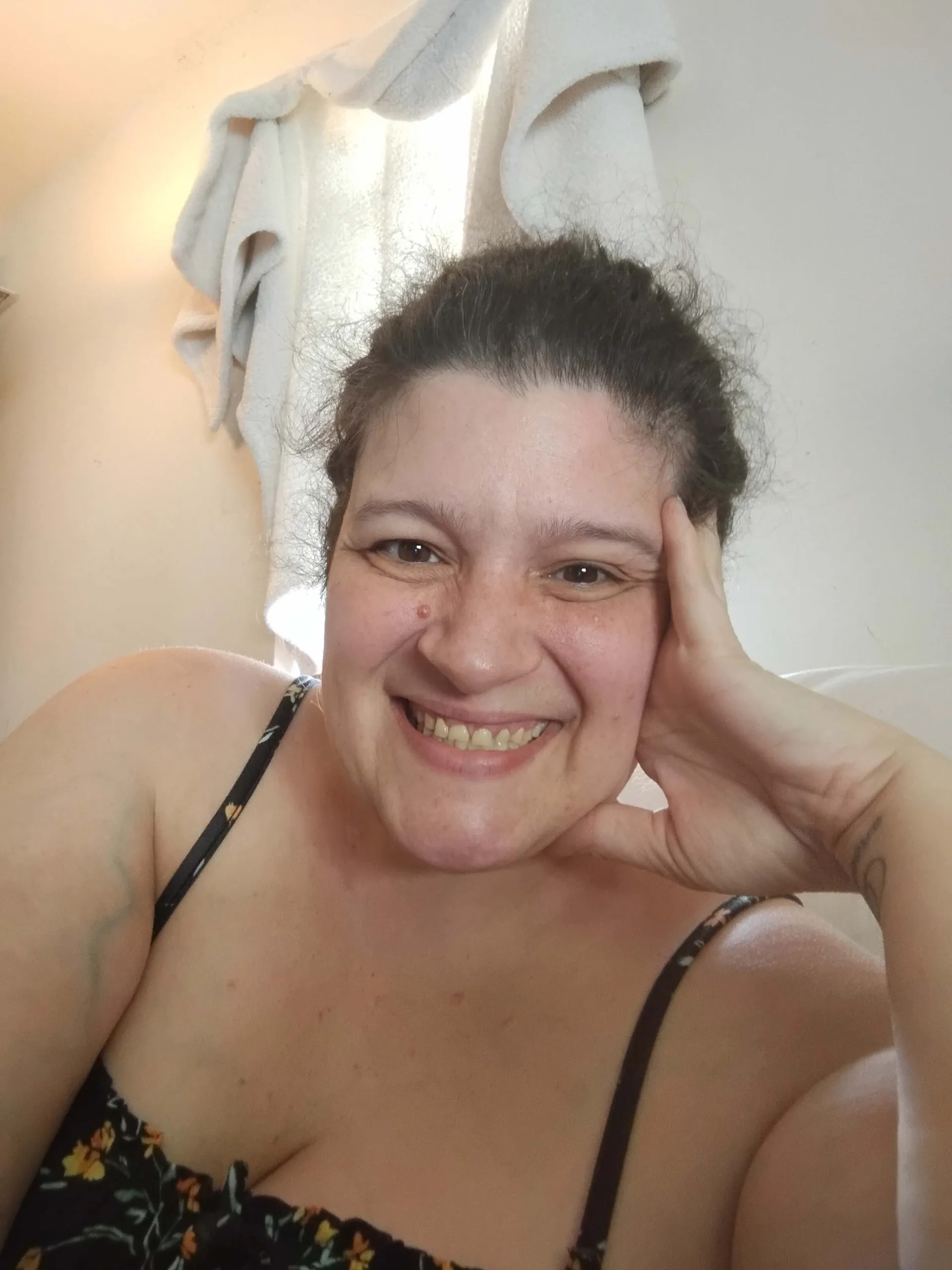 A close-up selfie of a person smiling with their hand resting on their cheek. They are wearing a floral print top.