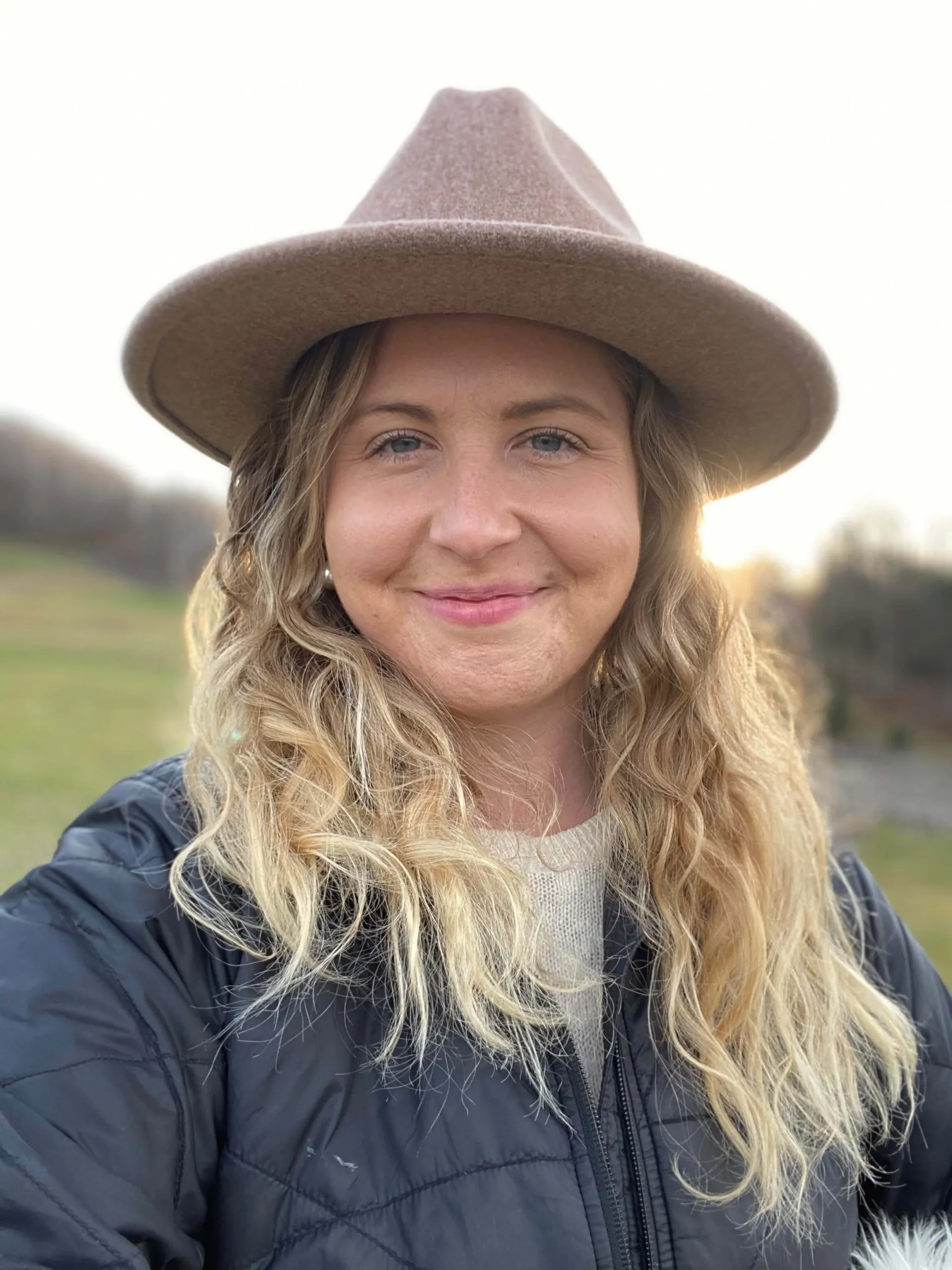 A person wearing a brown hat and a black puffer jacket smiles at the camera with long, wavy blonde hair. The background shows a blurred outdoor landscape with trees and a hint of sunset light.