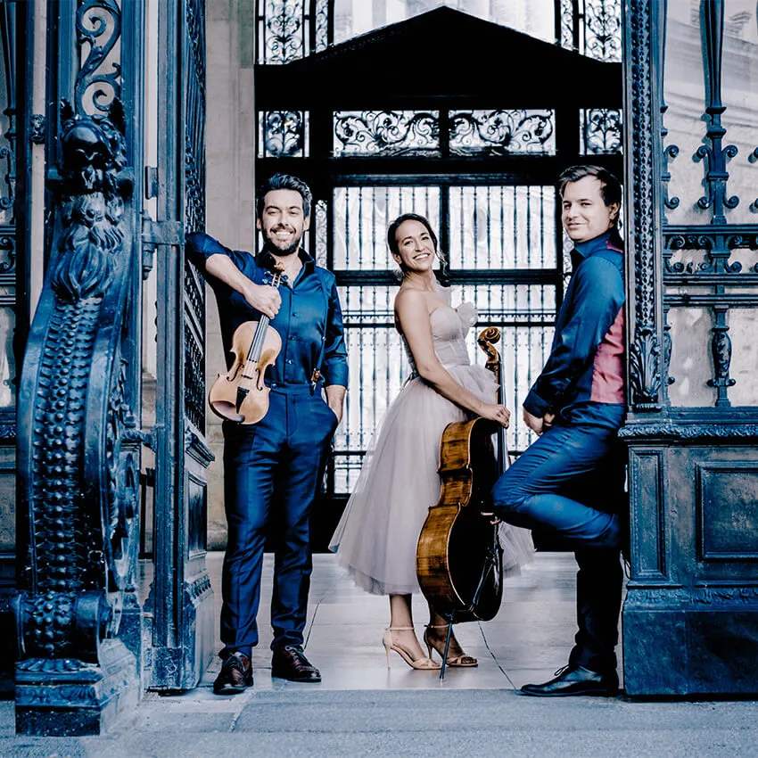 A woman and two men stand in a doorway, two of them holding classical instruments