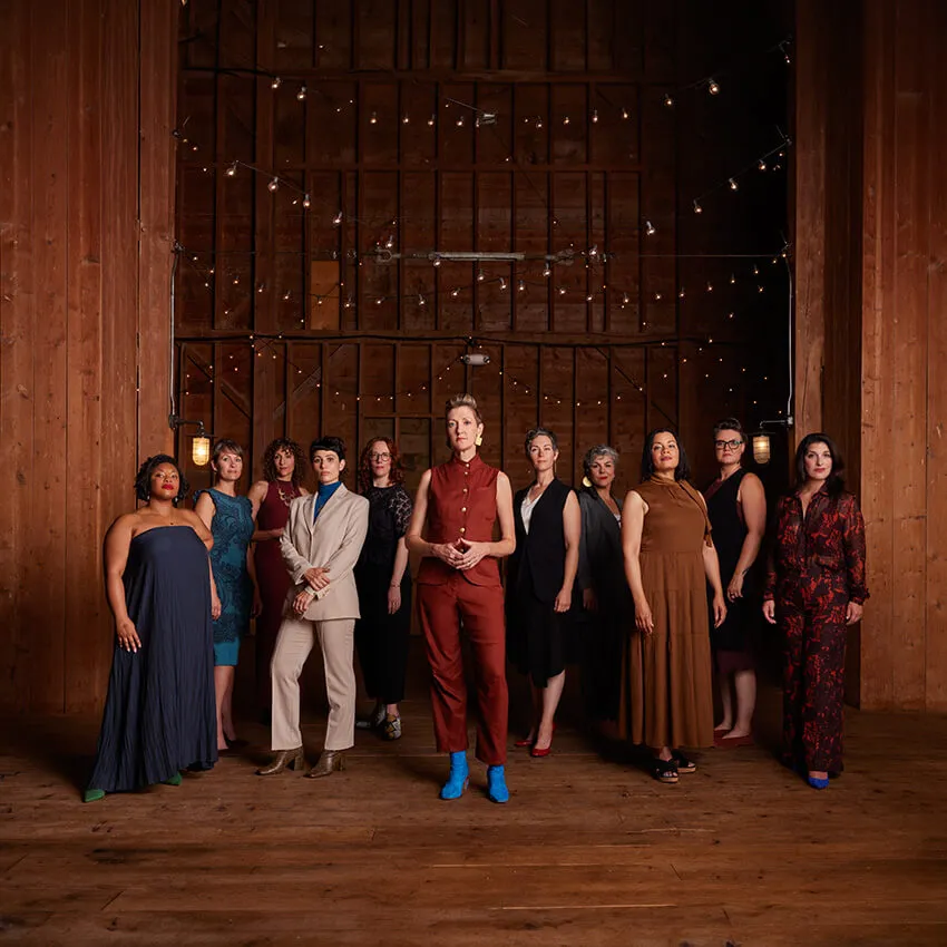 11 women stand in front of a large wooden door with string lights above them