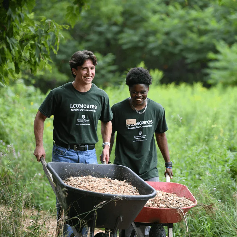 Two people outdoors pushing wheelbarrows full of woodchips smiling.