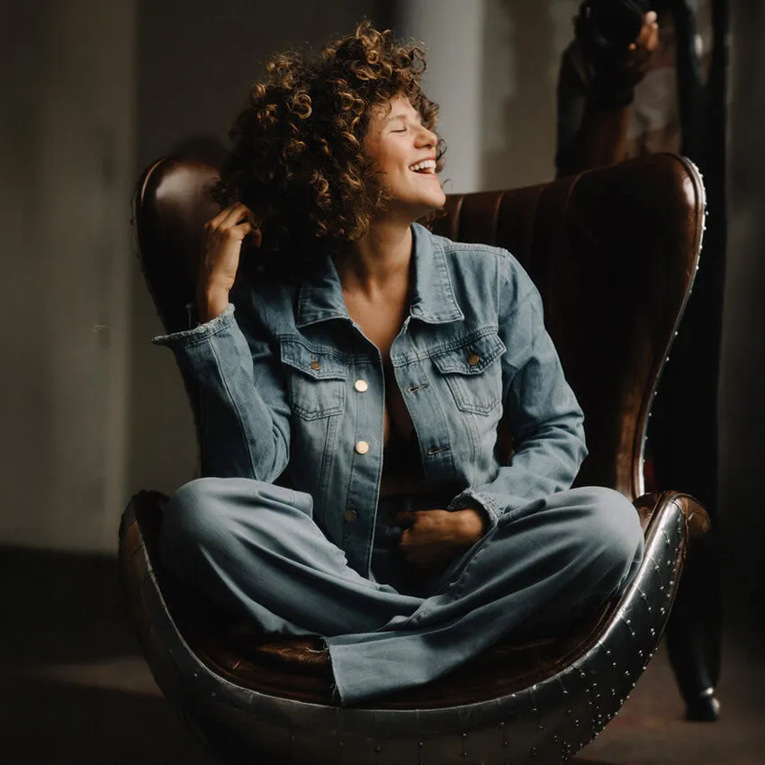 A woman with curly hair wearing jeans and a denim jacket sitting in a chair