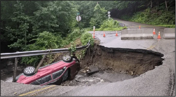 A photo showing a collapsed culvert that damaged a road during heavy rain, there is a red car in the large collapsed section of the road.