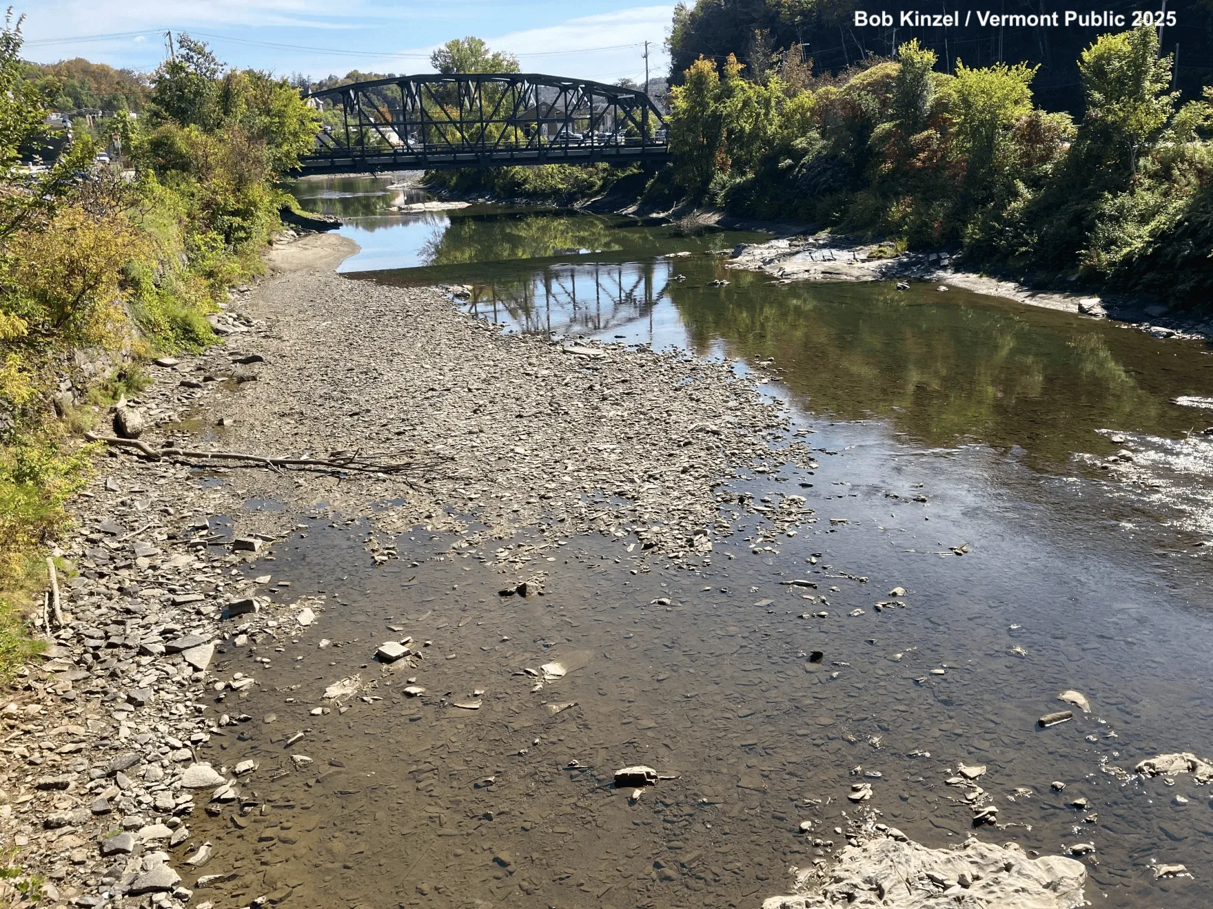 Photograph of the Winooski River. The water level is very low and the stream bed is visible with many small rocks. Green bushes and trees line the shore with a bridge in the background.